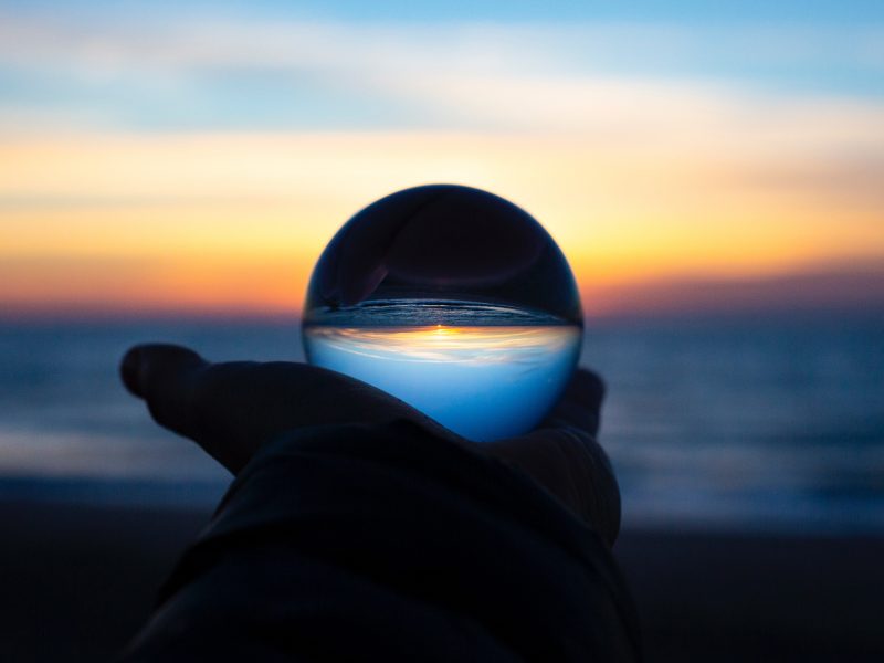 hand holding a clear glass ball in front of the sea. The sea and the sky are reflected on the ball, appearing upside down