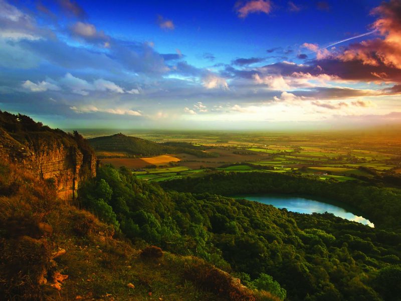Photo of a landscape, taken from the top of a mountain. There is a lake in the distance, woodlands, blue sky and sunset