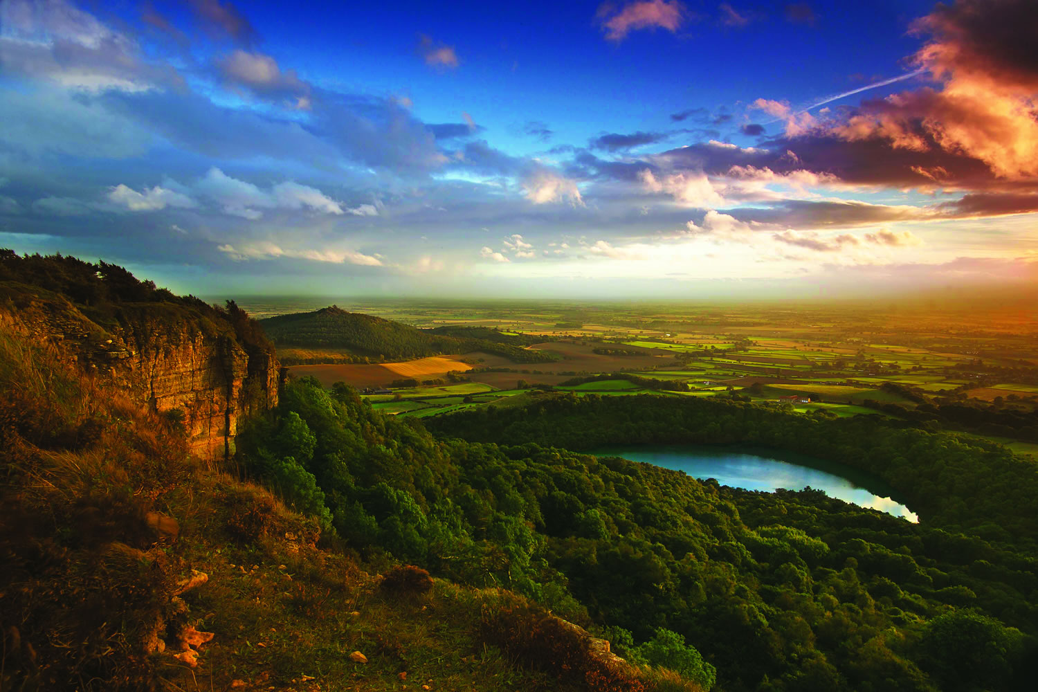 Photo of a landscape, taken from the top of a mountain. There is a lake in the distance, woodlands, blue sky and sunset