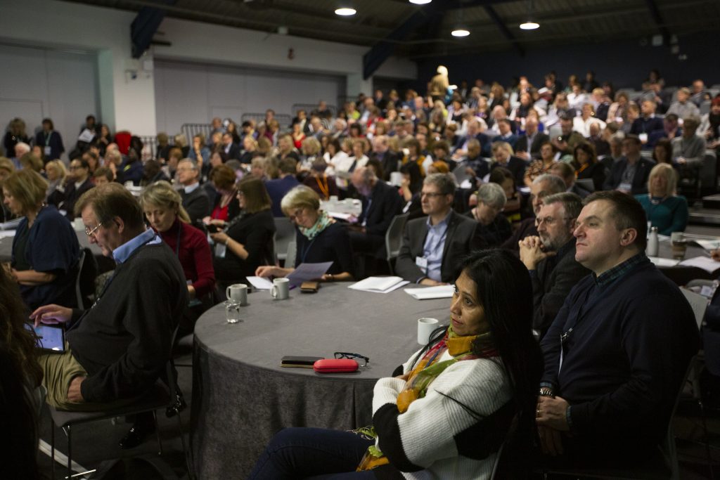 A large conference room filled with people sat listening to a speaker