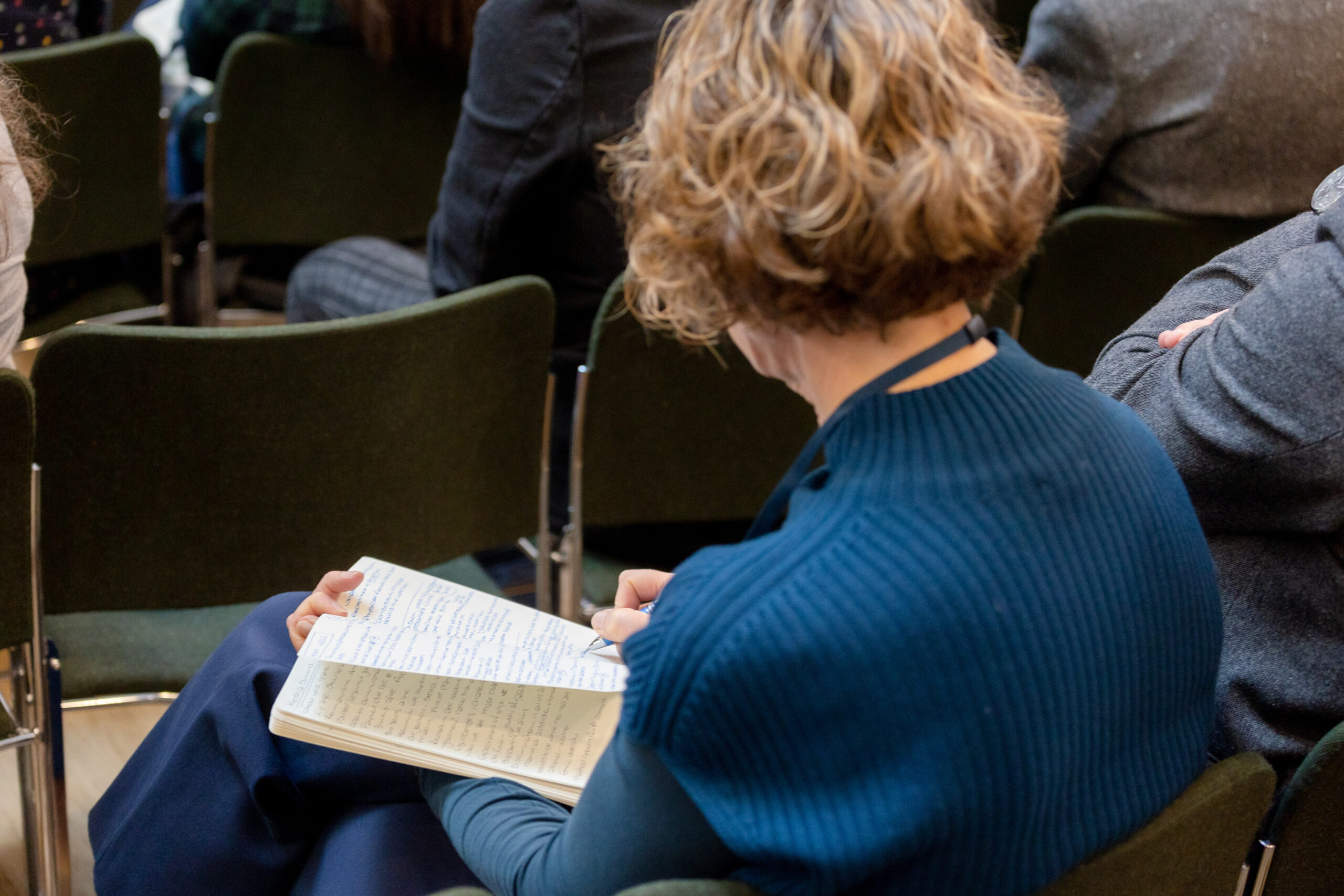 woman sitting down in an auditorium and taking notes on a notebook