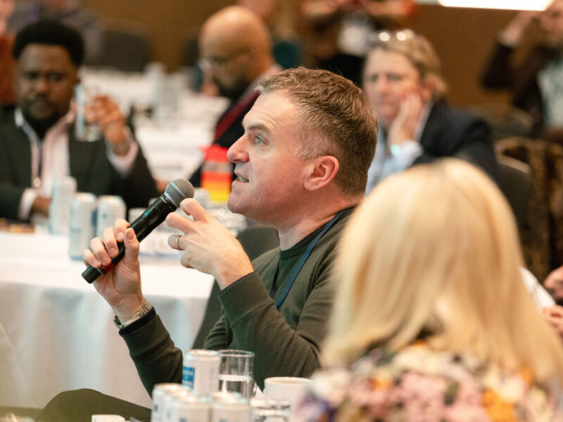 a man is holding a microphone, he is saying something while sitting among other people in a conference area