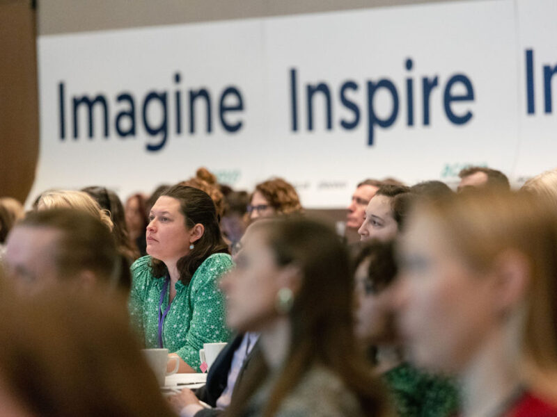 A photo of people sat at conference room tables listening to a speaker. The slogan imagine, inspire, improve is on the wall behind them