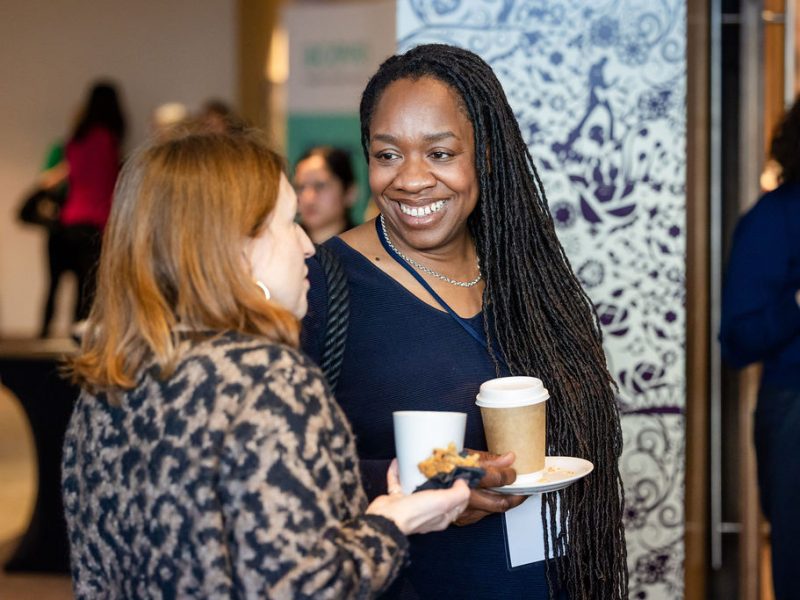 Two women stood in conversation, one is smiling at the other and they are holding coffee cups